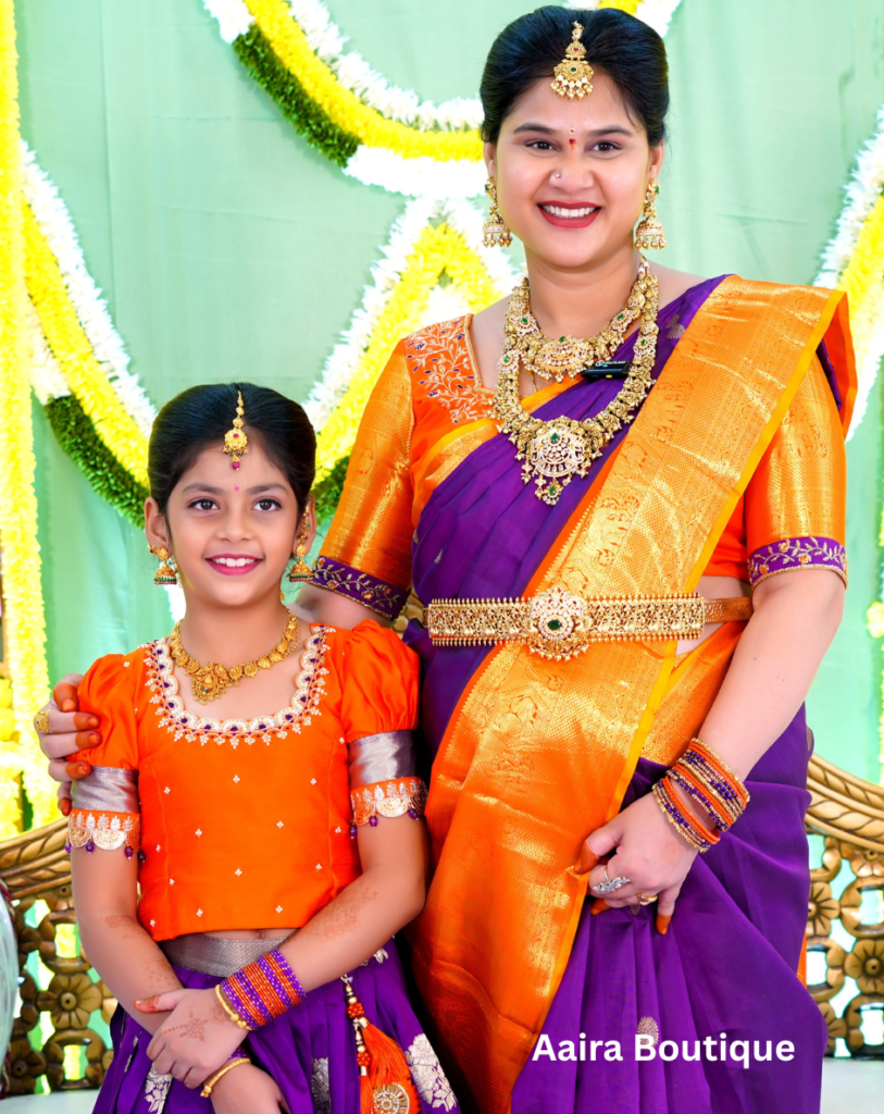 A smiling mother and daughter, both dressed in vibrant purple and orange traditional Indian ethnic wear, adorned with gold jewelry, standing in front of a festive yellow and green decorated backdrop.