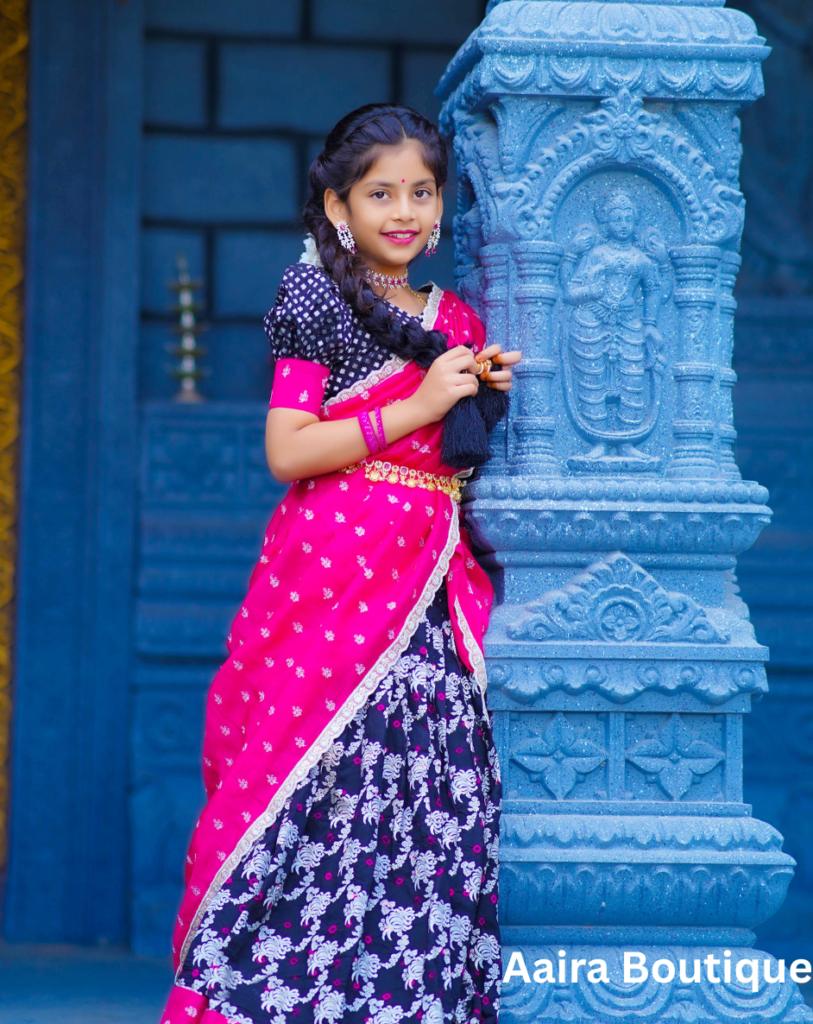 A young girl in a vibrant pink and black traditional Indian ethnic wear lehenga and matching blouse, with braided hair, standing by a carved blue pillar.