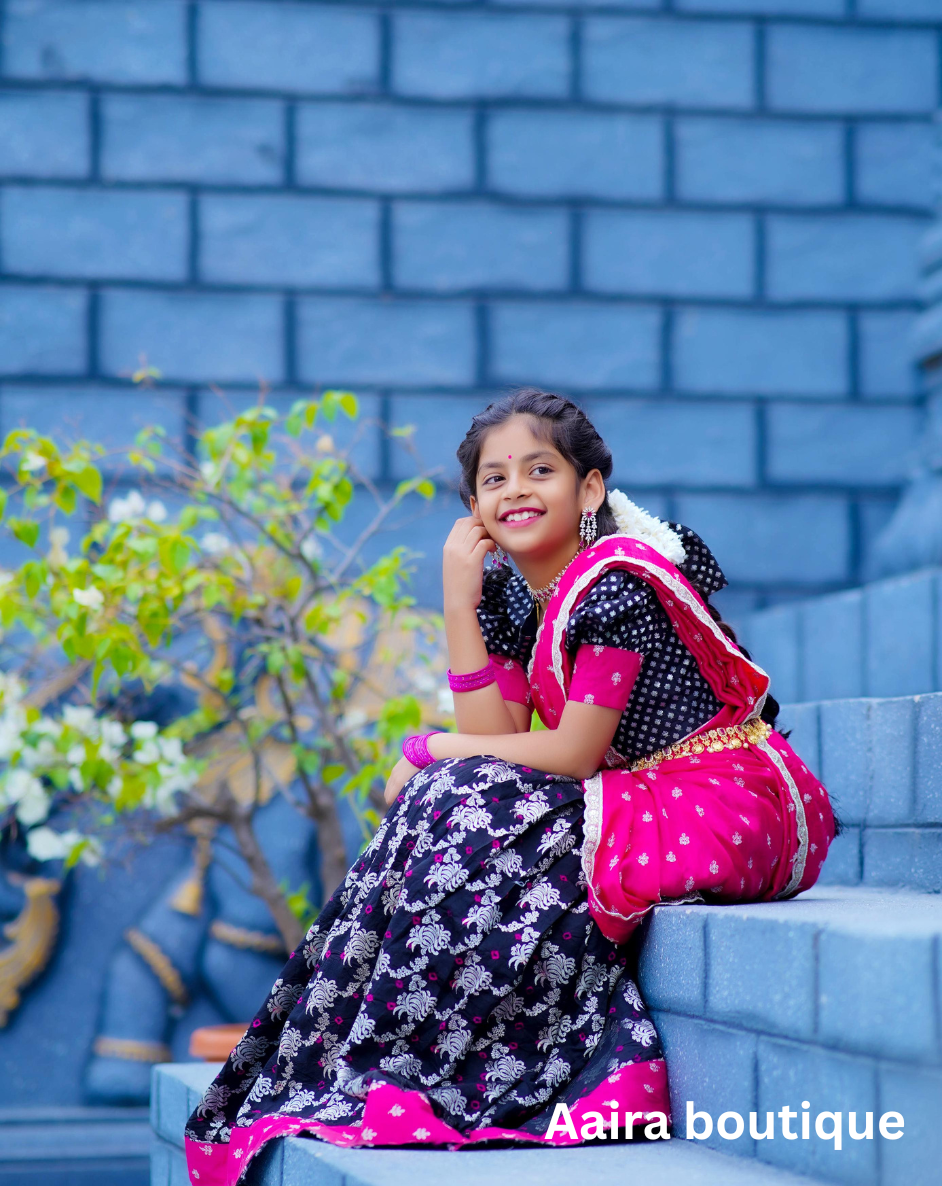 A young girl in a vibrant pink and black traditional Indian ethnic wear lehenga and matching blouse, with braided hair, standing by a carved blue pillar.